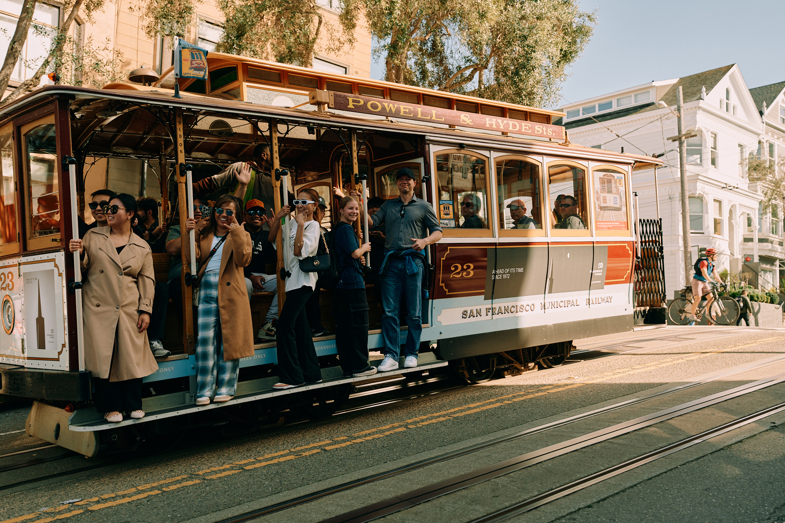 Tourists smiling and waving while riding San Francisco cable car.