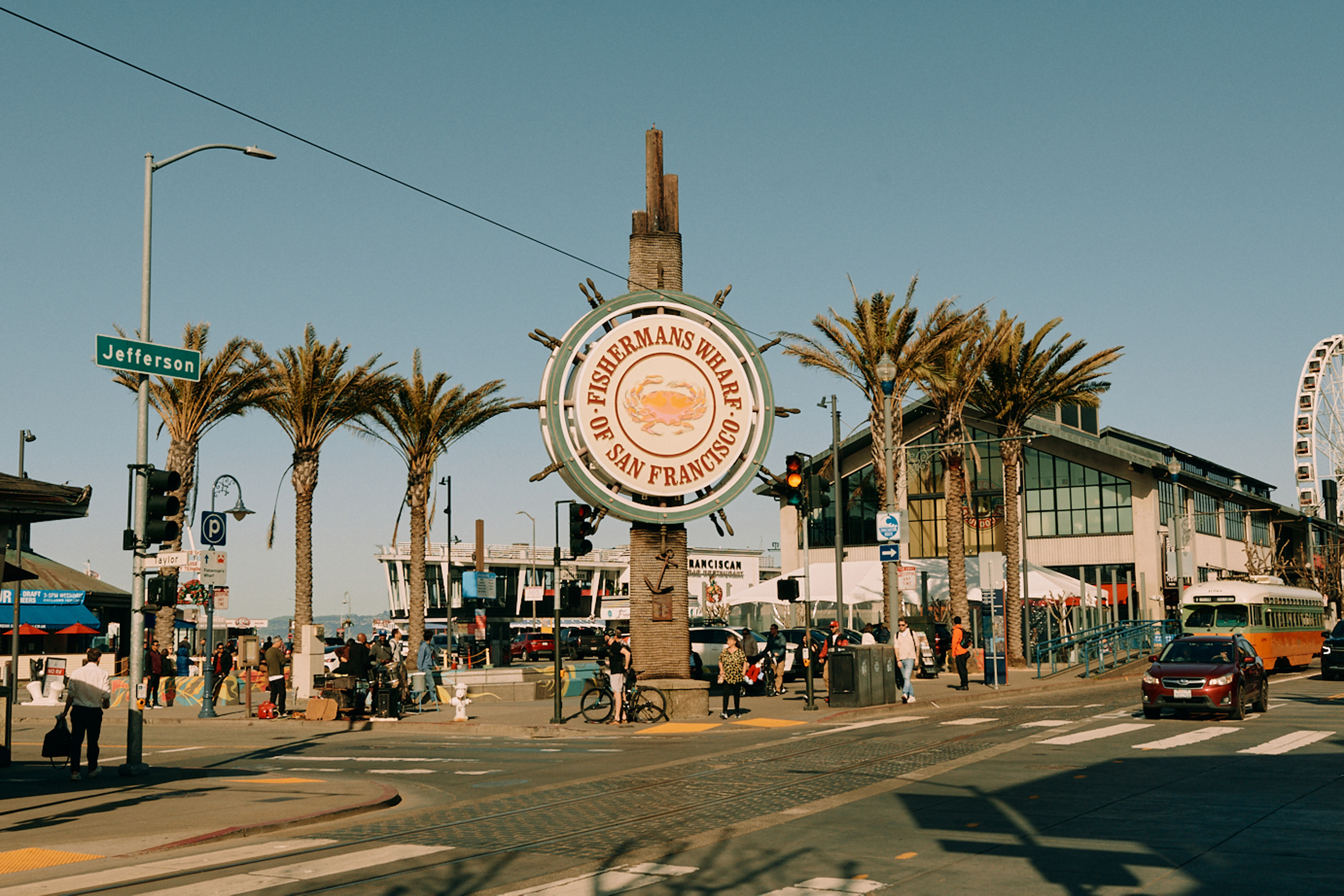 Fisherman’s Wharf sign surrounded by palm trees and street activity.